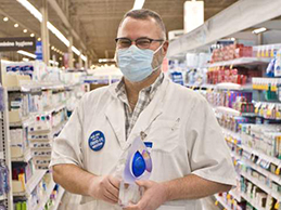 Kerry wears his white pharmacy coat and grins from behind his face mask as he holds his President’s Award trophy in the pharmacy aisle of his store.